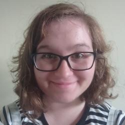 Photo of Cat Salvini. She is a white woman with brown/reddish shoulder length curly hair. She wears glasses, a black and white striped tshirt and blue dungarees.